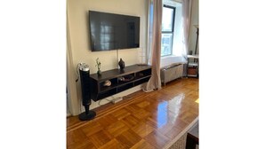 A living room with a flat-screen TV mounted on the wall, a dark wooden entertainment unit, a radiator, and a window with curtains. - PLG Boho Stay (Flatbush) (Brooklyn)