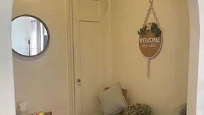 A cozy entryway with a wooden floor, a round mirror, a wicker chair with cushions, a small table with a plant, and a welcome sign on the wall. - PLG Boho Stay (Flatbush) (Brooklyn)