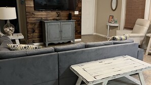 Ein Wohnzimmer mit einem grauen Ecksofa, einem weißen Couchtisch, einer Holzwand als Blickfang, einem Flachbildfernseher und einem Deckenventilator.