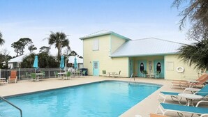 Ein Swimmingpool mit Liegestühlen und Sonnenschirmen, ein kleines Gebäude mit hellblauer Fassade und Palmen im Hintergrund.