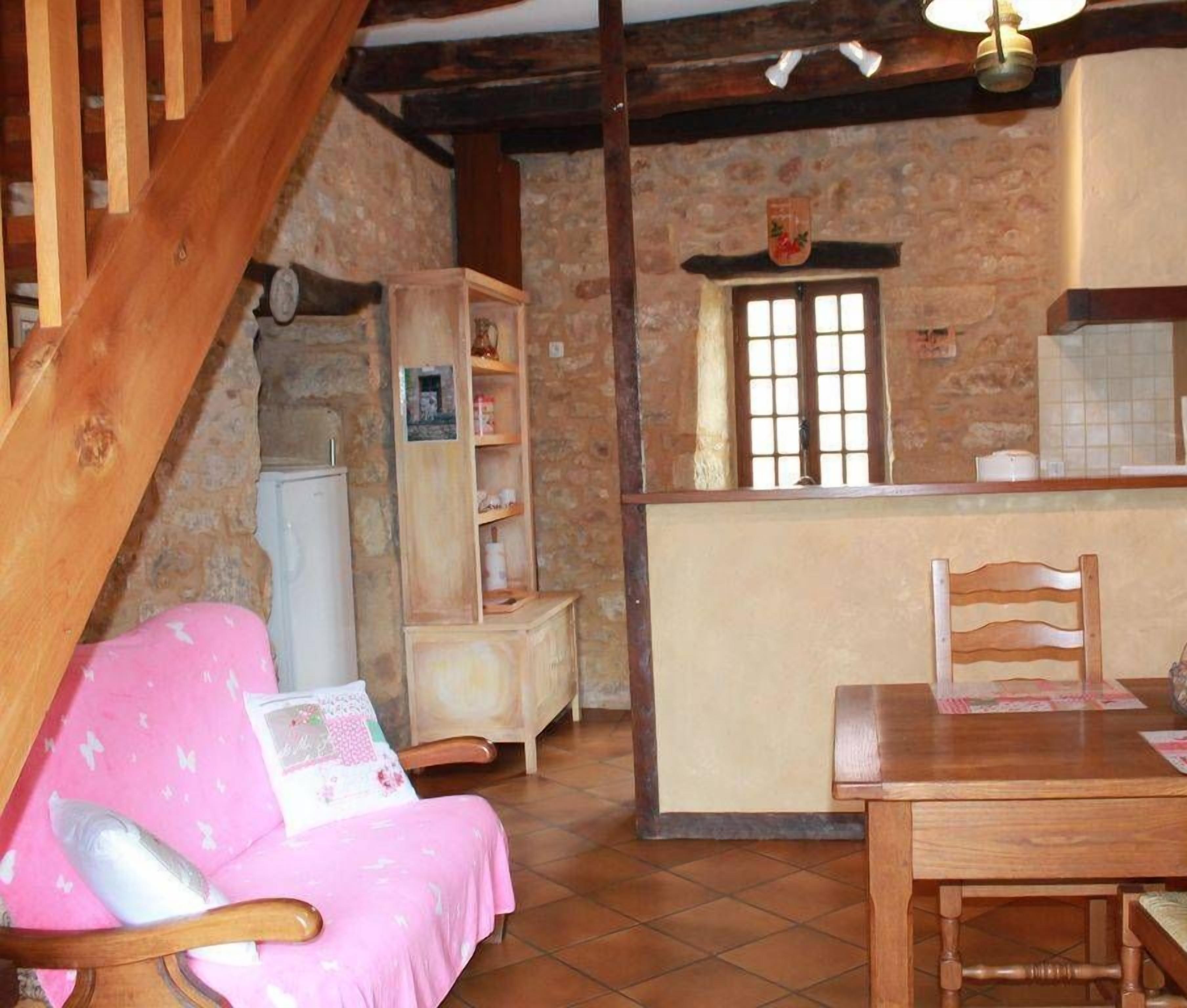 A cozy kitchen with a wooden dining table, a chair, and a pink chair with a pillow. A stone wall and a window are visible.