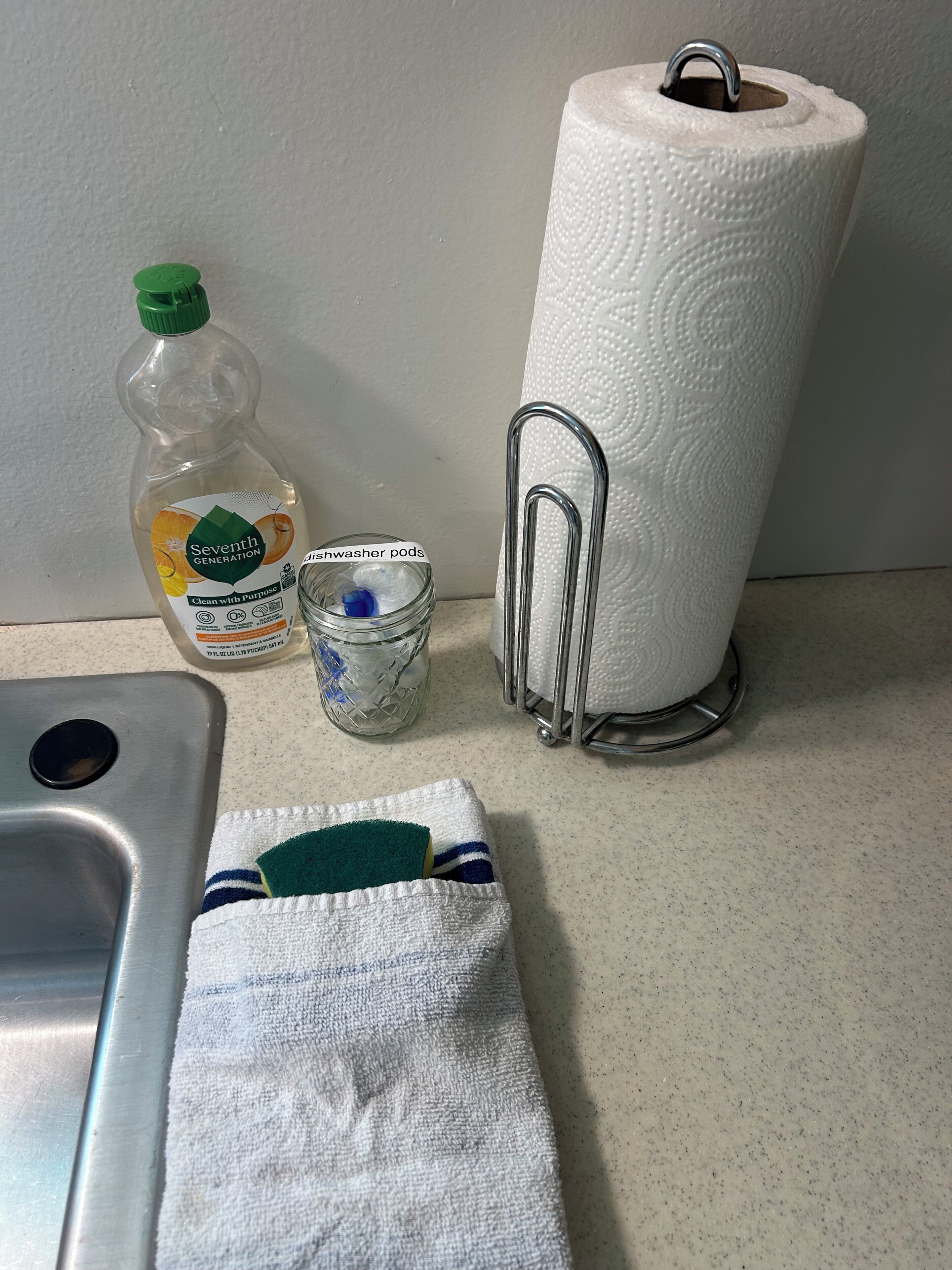 Una encimera de cocina con una botella de jabón para platos Seventh Generation, un frasco de vidrio con tapa azul, un dispensador de toallas de papel y una toalla.
