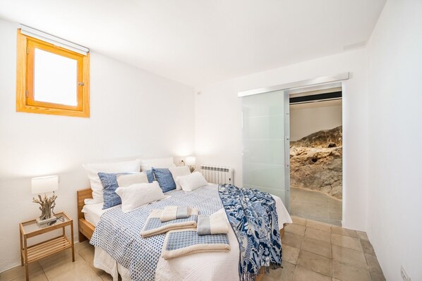 A bedroom with a bed, bedside table, lamp, and a view of a rocky outdoor area through a sliding door.