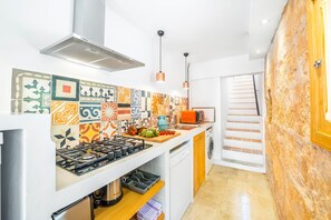 Una cocina moderna con estufa a gas, microondas y salpicadero de colores. Hay una encimera de madera con tabla de cortar y verduras. Se ve una lavadora en la esquina.