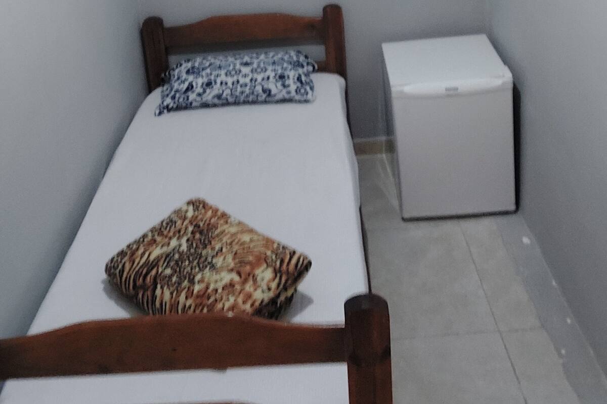 A small, narrow room with a single bed, a wooden headboard, a patterned pillow, and a white chest of drawers.
