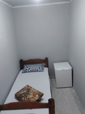 Une petite pièce étroite, avec un lit simple, une tête de lit en bois, un oreiller à motifs et une commode blanche.