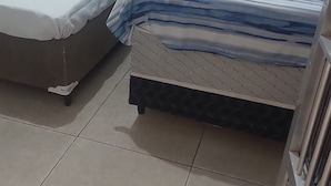 A small, compact room with two beds, one with a blue and white striped bedspread and the other with a plain white mattress. The room has white tiled walls and a tiled floor. - leandrohoteis23 (Guarulhos)