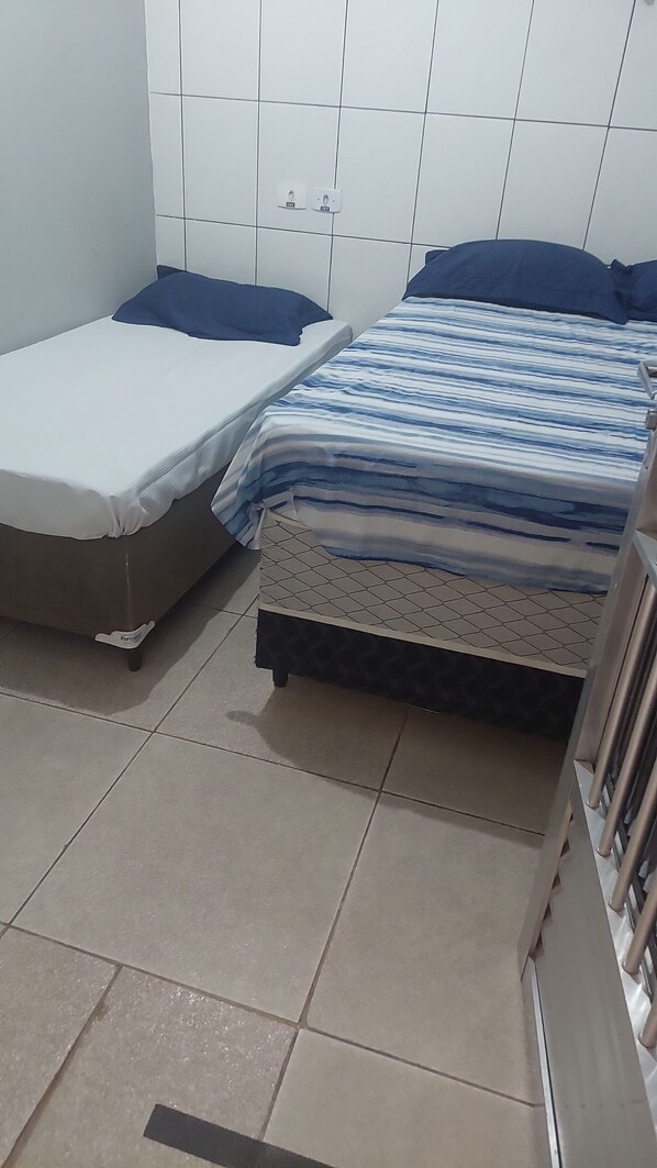 A small, compact room with two beds, one with a blue and white striped bedspread and the other with a plain white mattress. The room has white tiled walls and a tiled floor.