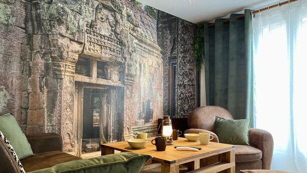 Een gezellige woonkamer met een muurschildering van een stenen muur, een houten salontafel met kopjes en een lantaarn, en een bruine leren bank.