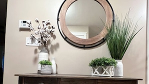 Une console en bois foncé, surmontée d’un miroir rond, agrémentée de deux vases avec des plantes et d’une petite étagère avec des coussins.