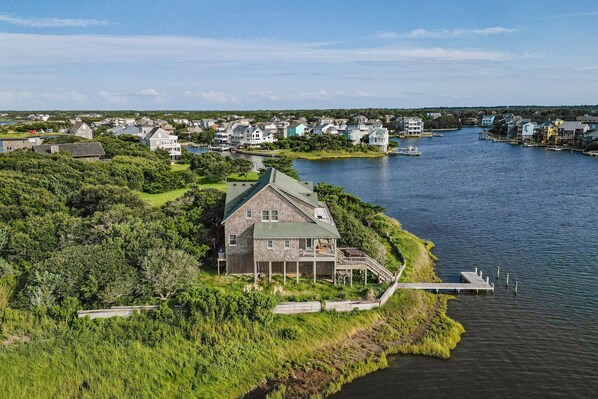 A large house with a green roof and a dock extending into a lake, surrounded by other houses and greenery.