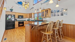 A kitchen with wooden cabinets, a black refrigerator, a wooden island with bar stools, and wall-mounted lights. - 7825-The Carolina Moon - Soundfront Frisco Home | Sunset Views (Frisco)