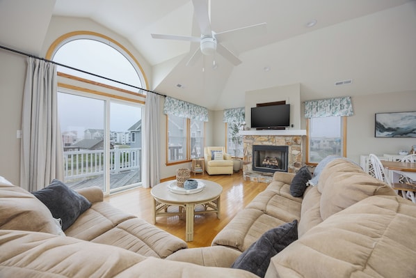 A spacious living room with a high ceiling, a fireplace, a television, and a balcony with a view of houses.