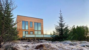 Una casa moderna con amplios ventanales y fachada de madera, ubicada junto a una masa de agua tranquila con vegetación circundante.