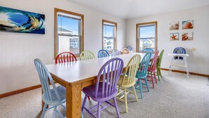A dining room with a long table and colorful chairs, large windows, and a beach-themed painting on the wall. - 5418- Family Tides - Semi-Oceanfront OBX Home for Large Groups (Nags Head)