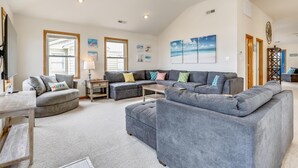 A spacious living room with a grey sectional sofa, a small table, and a lamp. There are large windows allowing natural light, and artwork on the walls. - 5418- Family Tides - Semi-Oceanfront OBX Home for Large Groups (Nags Head)