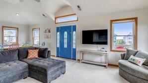 A modern living room with a sectional sofa, a grey ottoman, a TV, and a dining area with colorful chairs. - 5418- Family Tides - Semi-Oceanfront OBX Home for Large Groups (Nags Head)