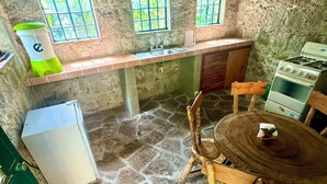 Una cocina rústica con suelo de piedra, sillas de madera, una mesa pequeña, un refrigerador, un lavadero y una estufa.