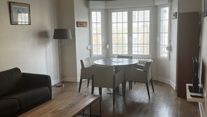 Un salon moderne avec une table basse en bois, un canapé noir, une table de salle à manger ronde avec des chaises, un lampadaire et une grande fenêtre donnant sur l’extérieur.