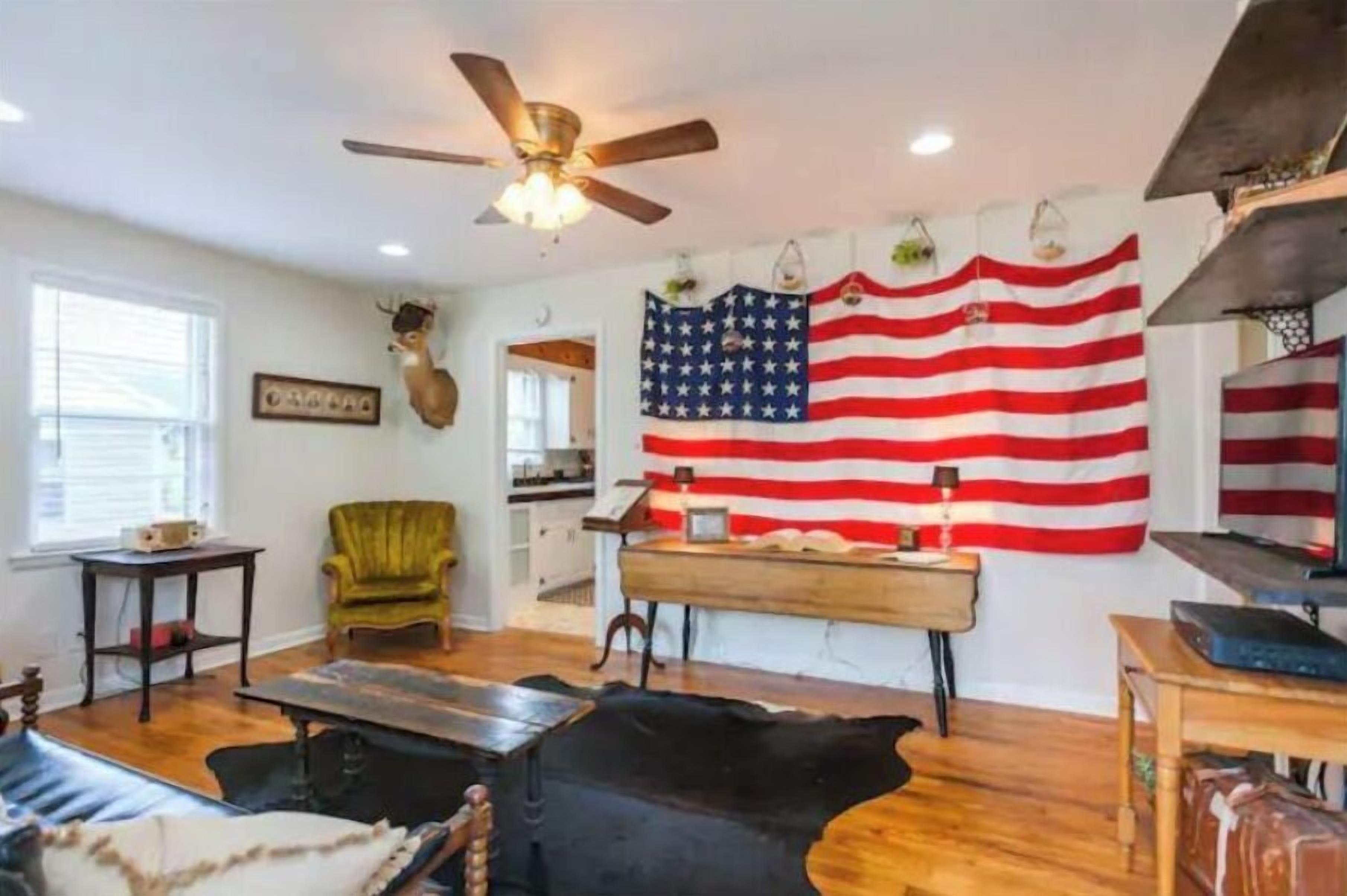 A living room with a ceiling fan, a large American flag, a deer head decoration, and a green armchair.