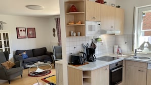Eine kompakte Küche mit Mikrowelle, Kaffeemaschine und Backofen. Im Vordergrund steht ein Esstisch mit einer Vase getrockneter Pflanzen.