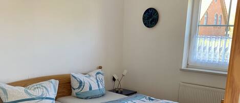 A bedroom with a wooden bed, a bedside table with two lamps, a wall clock, and a window with a view of buildings.