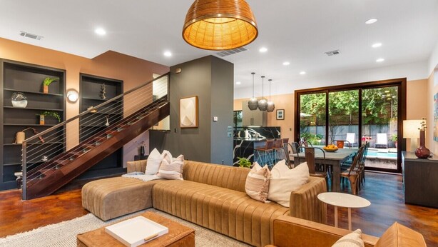 A modern living room with a sectional sofa, a wooden coffee table, a staircase, and a dining area with a view of the outdoors.