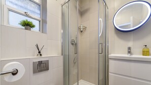 Ein modernes Badezimmer mit einer gläsernen Duschkabine, einem Toilettenpapierhalter, einem Waschbecken mit Seifenspender und einem Fenster mit Pflanze.