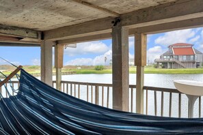 A hammock on a wooden deck overlooking a body of water with a house in the background.