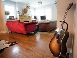 Un salon avec un canapé rouge, un divan en cuir marron, un plancher en bois, une tête de cerf accrochée au mur, une guitare et une cheminée.