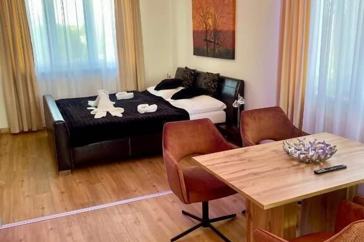 A modern hotel room with a bed, a wooden dining table, and two armchairs.