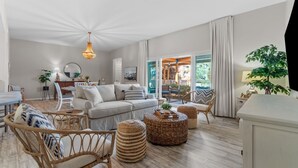 Un salon spacieux avec un canapé gris clair, des fauteuils en osier et une table basse ronde. Une grande fenêtre avec des rideaux, un luminaire suspendu et une vue sur l’extérieur complètent l’ensemble.