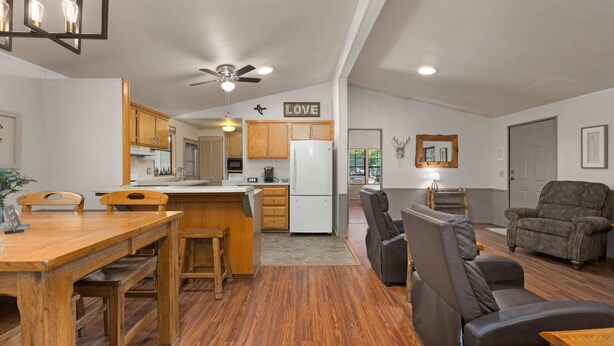A spacious living area with a wooden dining table, a sofa, and a recliner. A kitchen with wooden cabinets and a refrigerator is visible in the background.