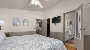 A bedroom with a large bed, a ceiling fan, a TV mounted on the wall, and a walk-in closet.