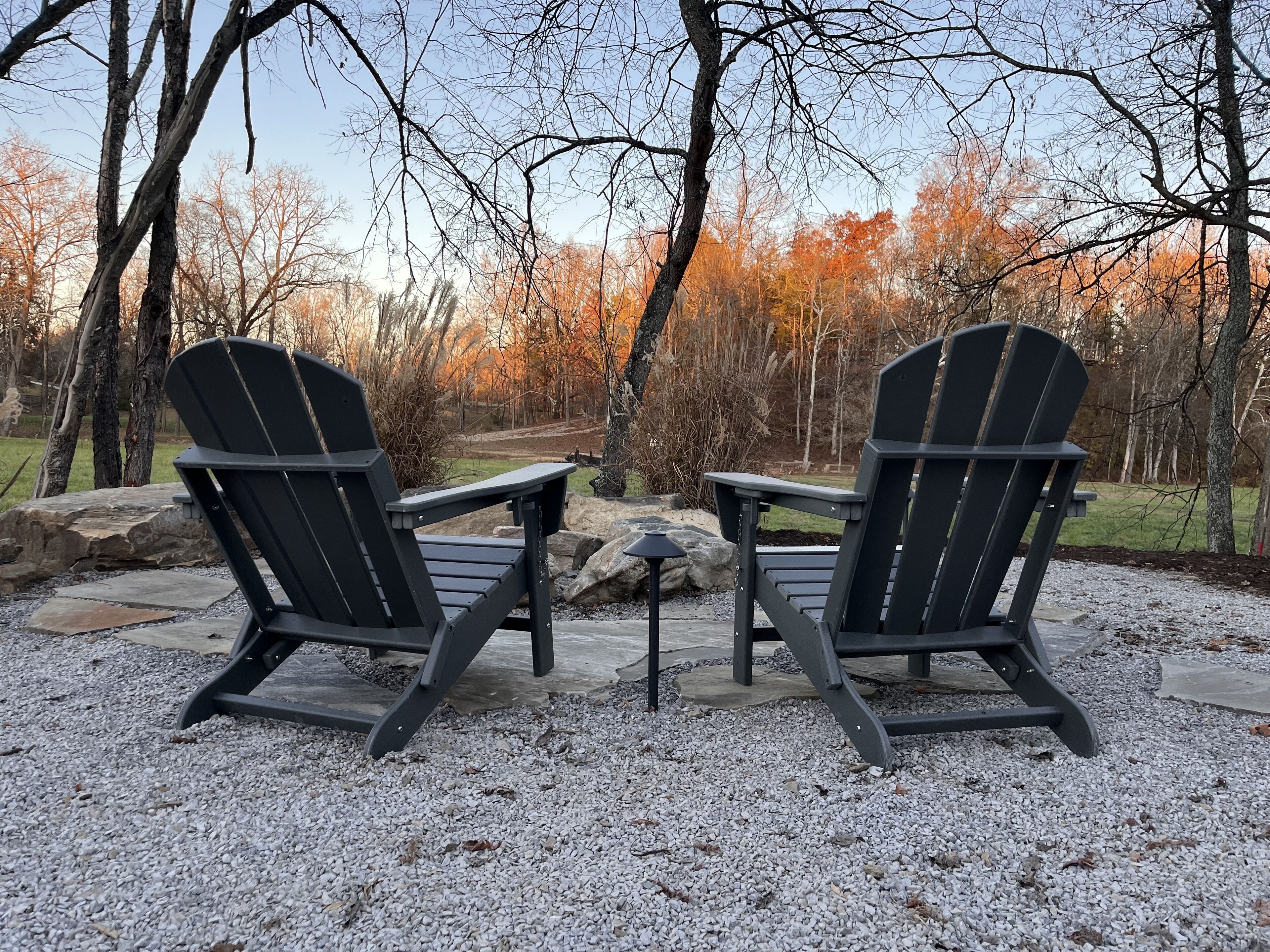 Duas cadeiras Adirondack viradas uma para a outra, com uma pequena mesa entre elas, dispostas sobre um piso de cascalho, com árvores ao fundo.