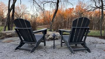 Duas cadeiras Adirondack viradas uma para a outra, com uma pequena mesa entre elas, dispostas sobre um piso de cascalho, com árvores ao fundo.