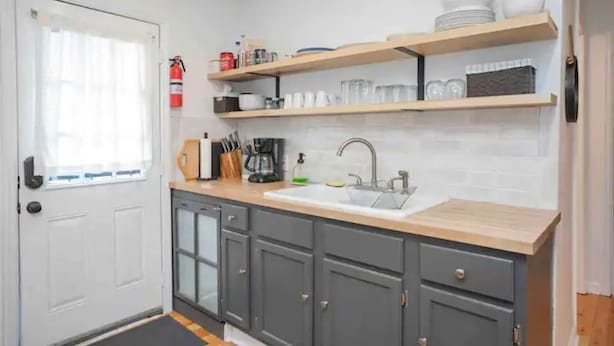 Una cocina compacta con armarios grises, encimera de madera y un lavabo blanco. Hay estantes sobre el lavabo con platos y vasos.