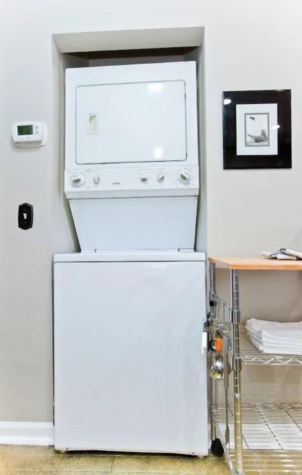 A compact laundry room with a front-loading washer and dryer, a small shelf with folded towels, and a framed picture on the wall. - The Ruby at Music Row (Nashville)