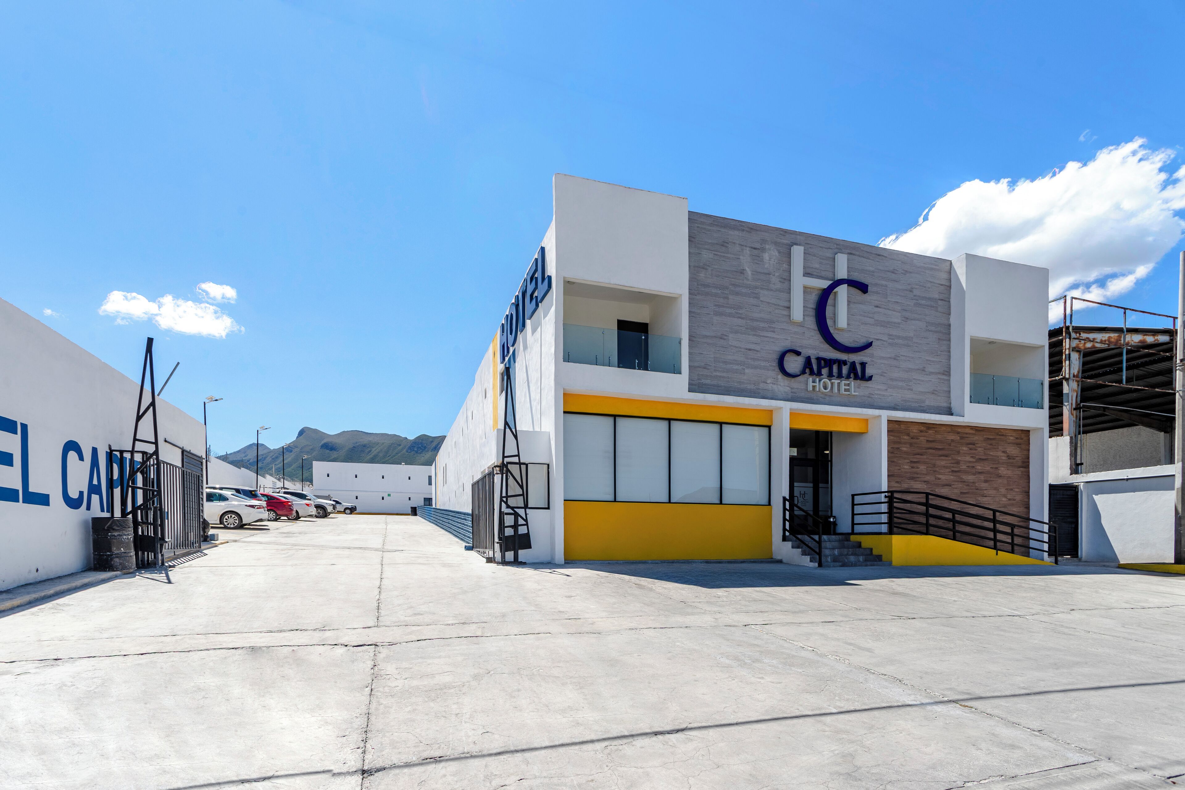 Un hotel moderno con un llamativo letrero que dice 'Capital Hotel', una zona de estacionamiento y montañas al fondo.