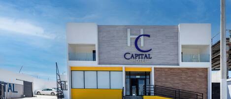 Un hotel moderno con un llamativo letrero que dice "Capital Hotel", una entrada con portón y un área de estacionamiento.