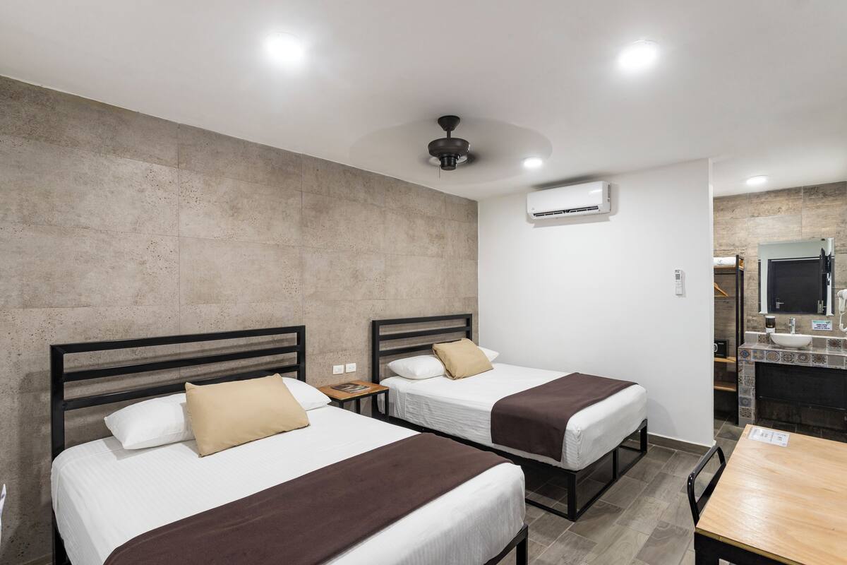 A hotel room with two beds, a wooden table, a bathroom with a sink and mirror, and a ceiling fan.