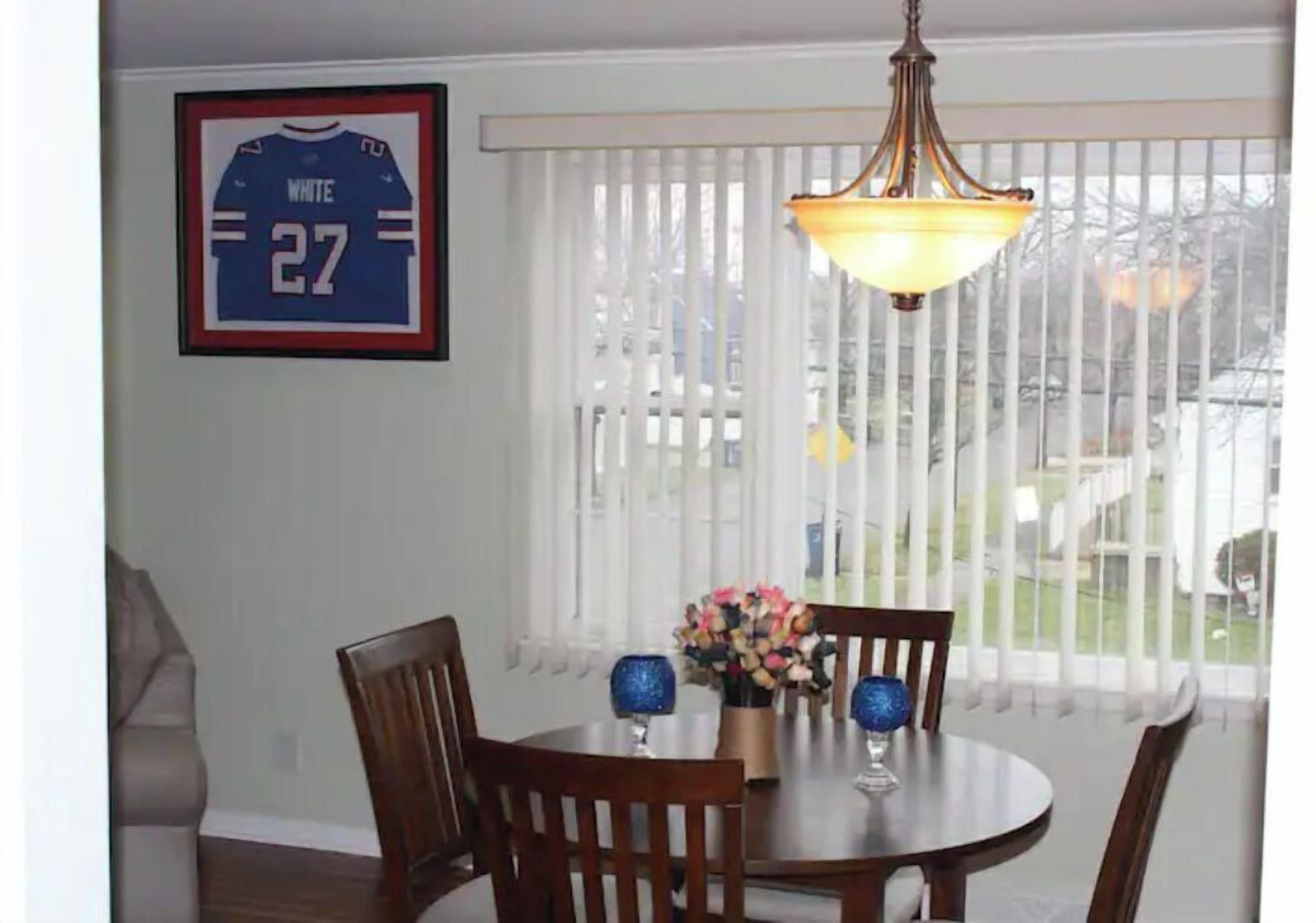 A dining area with a round table, chairs, a chandelier, a framed sports jersey, and a window with blinds.