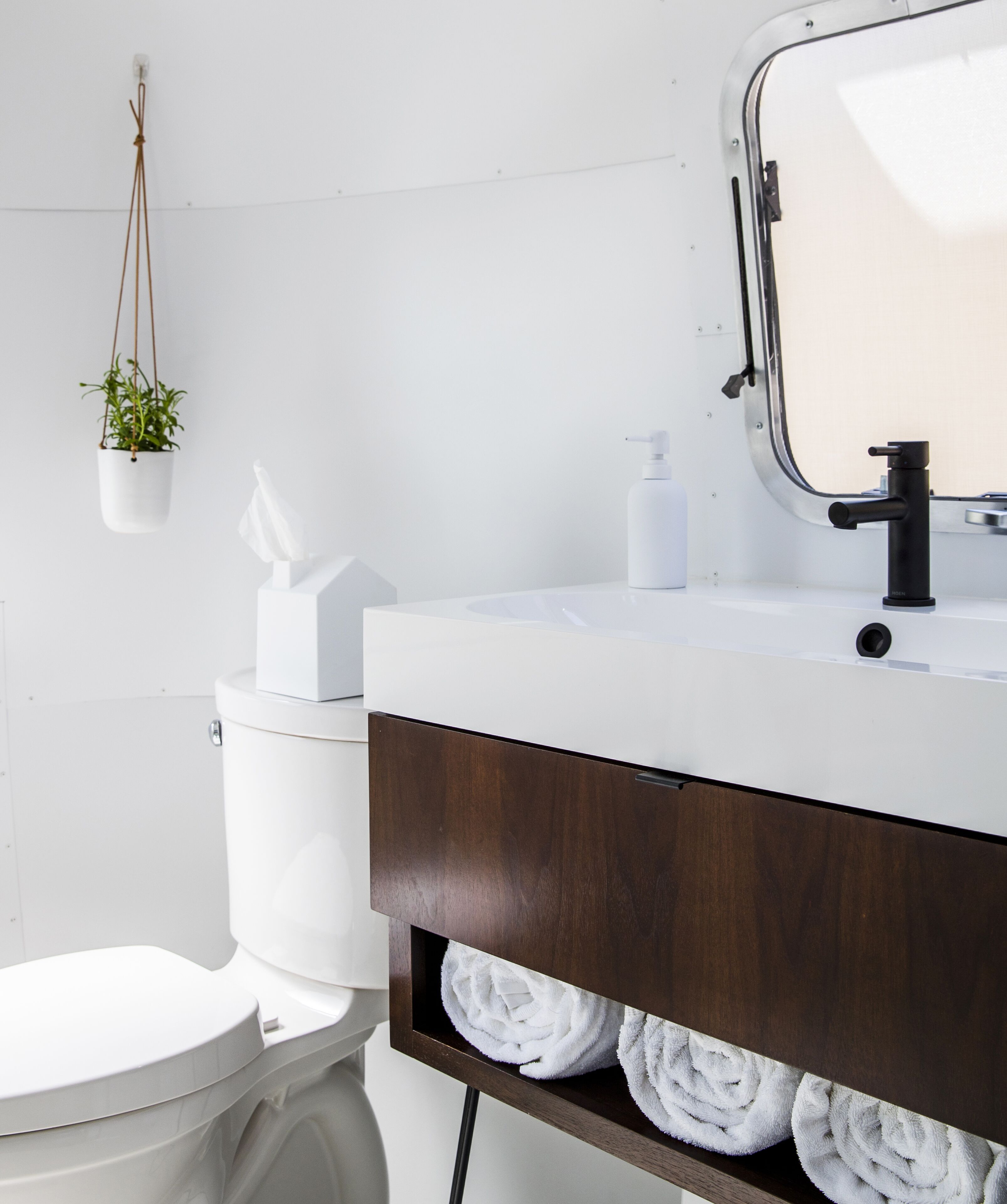 A modern bathroom with a white toilet, a white sink with a wooden vanity, a hanging plant, and a towel rack with towels.