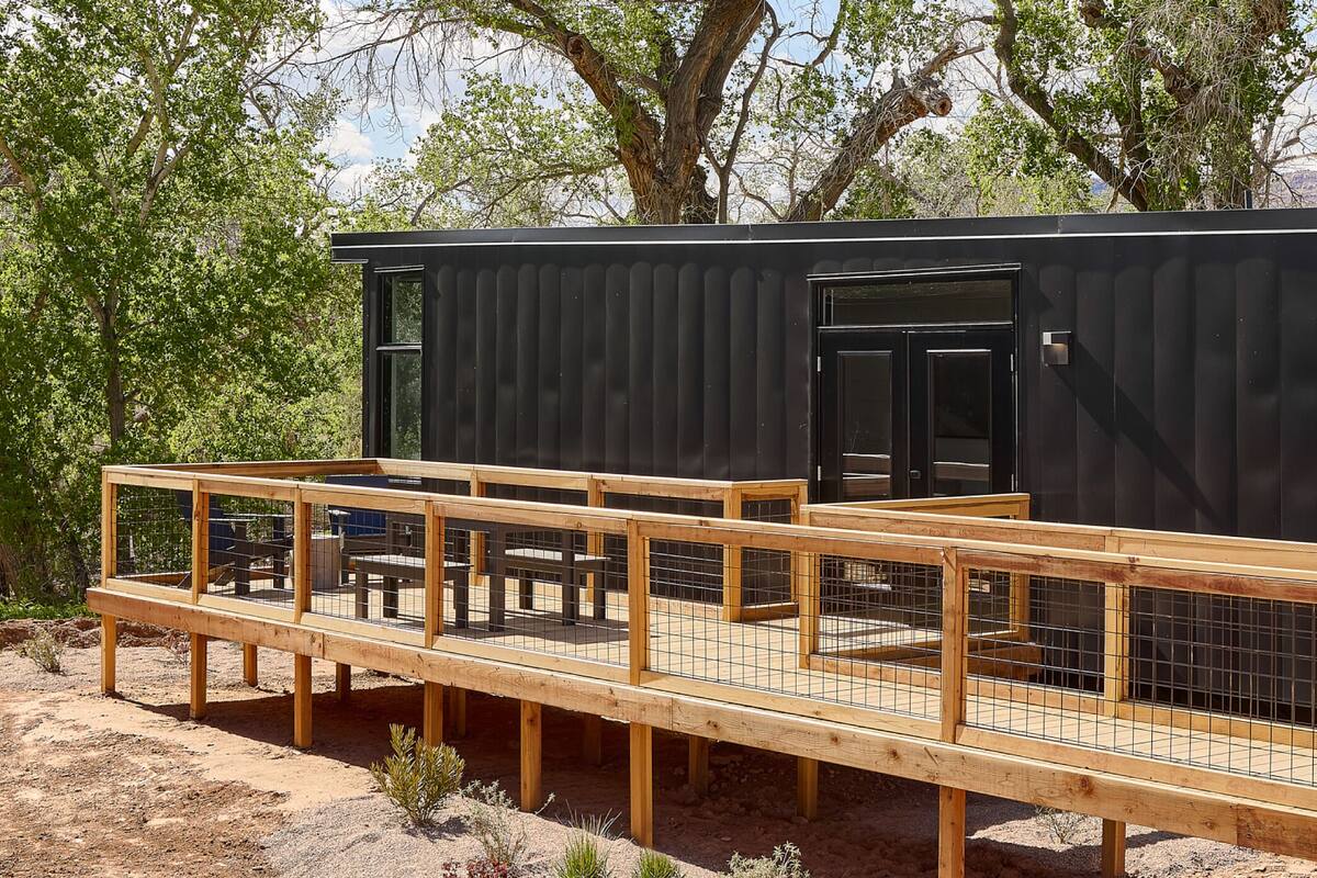 A modern cabin with a wooden deck and a black exterior, surrounded by trees.