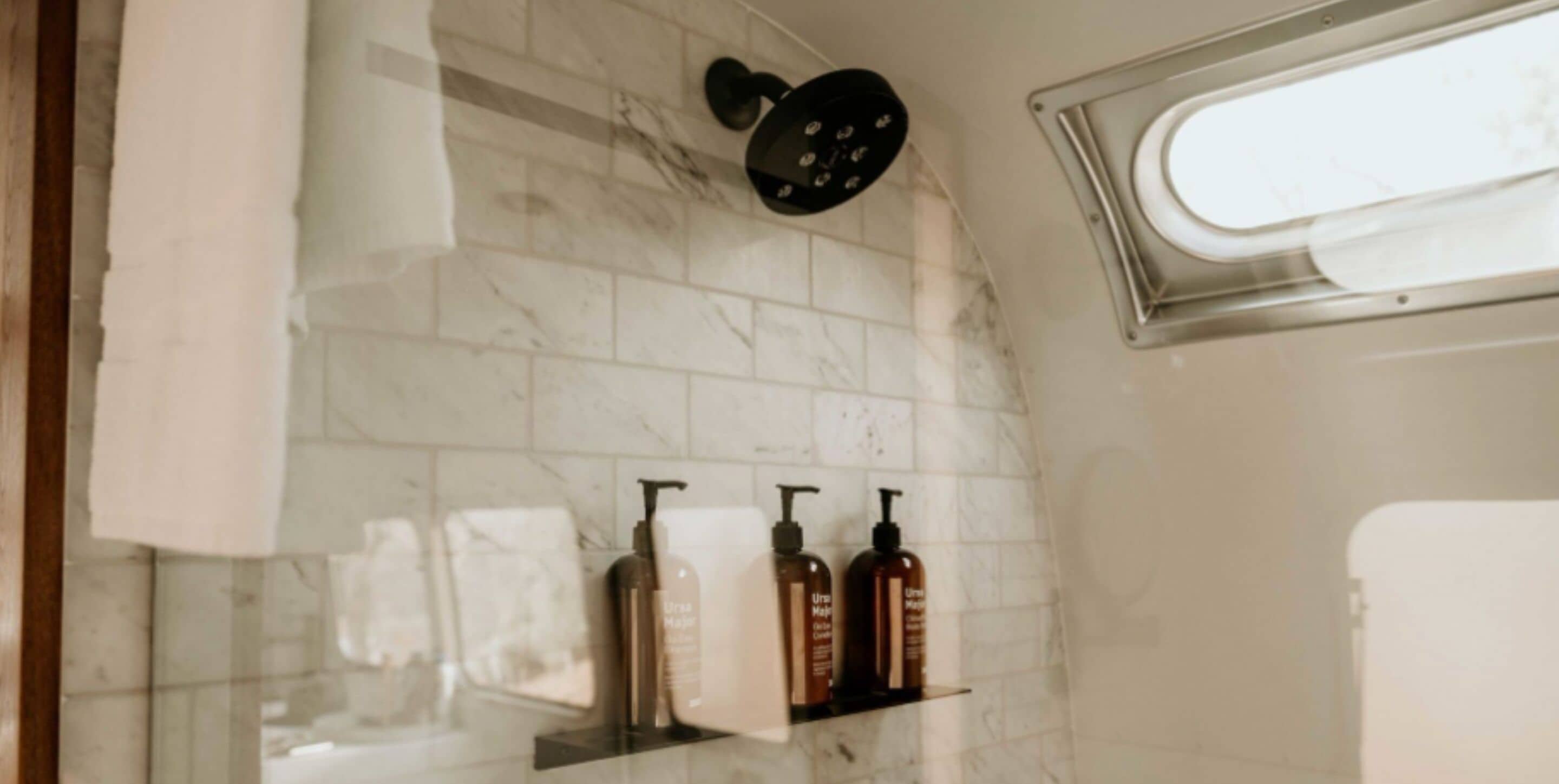 A bathroom with white marble tiles, a black showerhead, and three bottles of shampoo on a shelf.