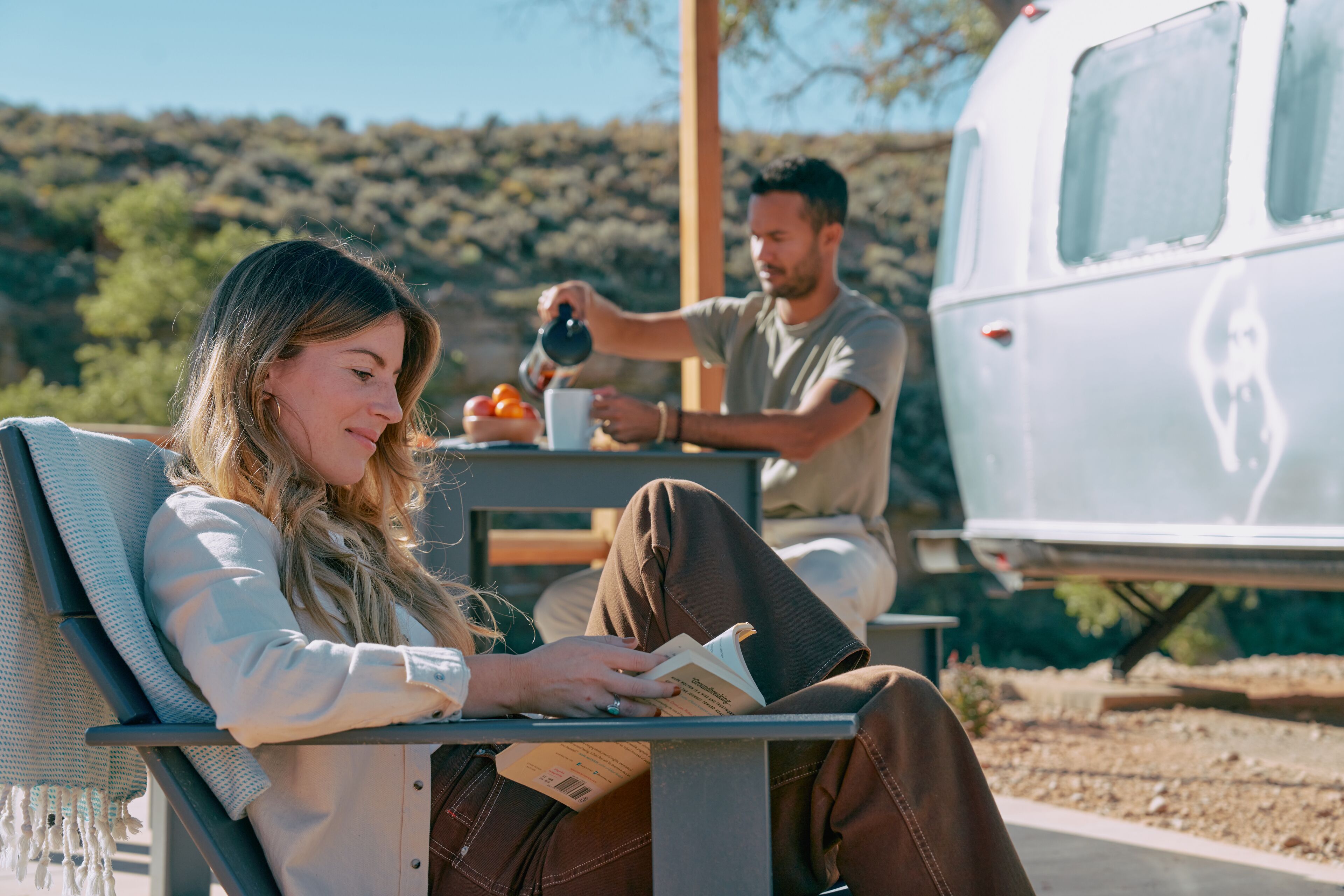 A person sitting on a bench reading a book, another person pouring a drink, and a white camper van in the background.