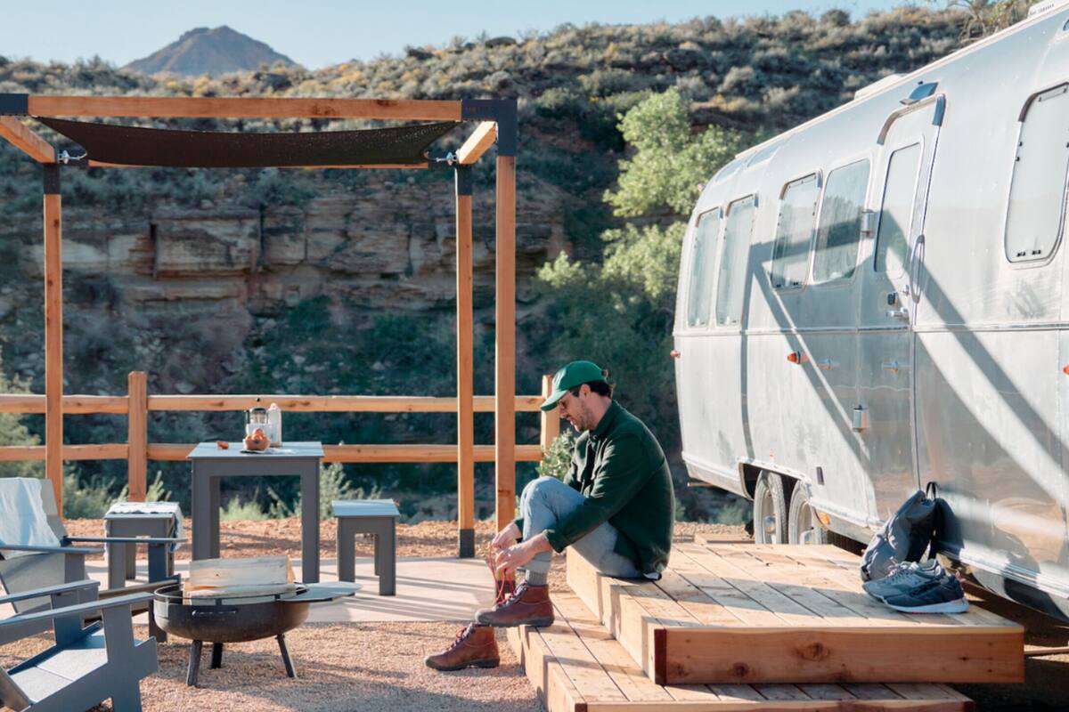 A campsite with a wooden table, chairs, a grill, and a camper parked in a desert-like environment with rocky hills in the background.