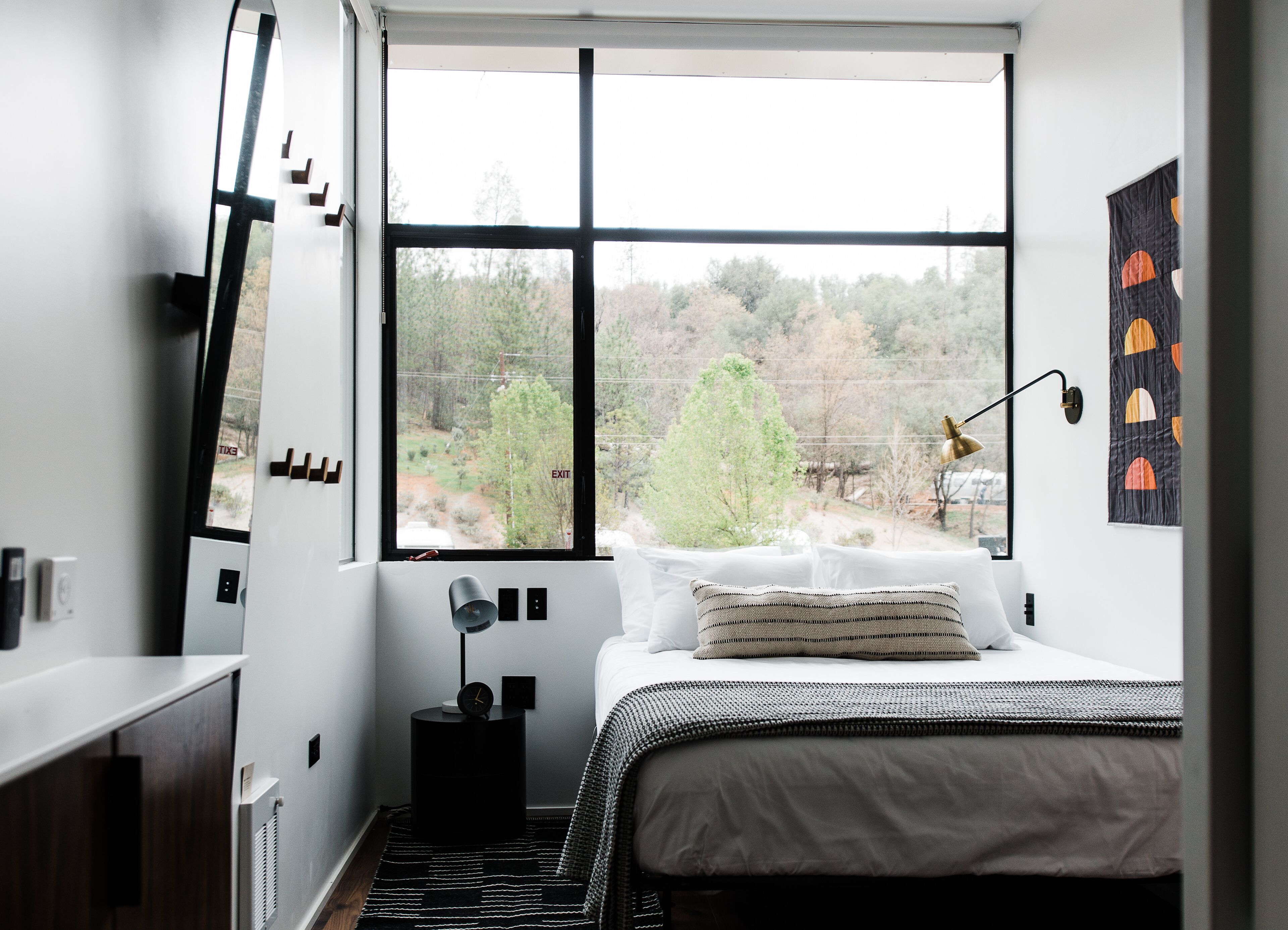 A modern bedroom with a large window, a bed with a striped pillow, a bedside lamp, and a small black side table with a clock.