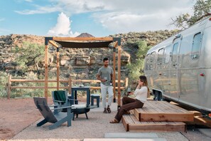 Een houten pergola met een tafel, stoelen, een bankje en een aanhanger, gelegen in een woestijnlandschap.