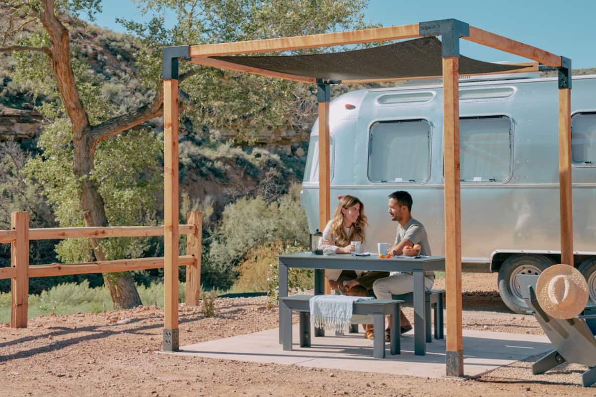 A couple dining at an outdoor table under a wooden pergola, with a vintage camper parked nearby.